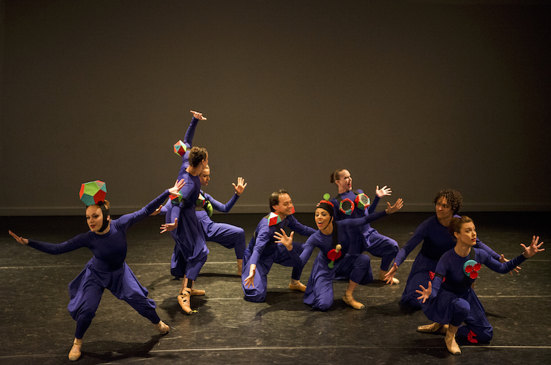 A large group of women in purple unitards with flowy pants. They each wear a cartoonish accessary like an oversized ring as a headpiece. 